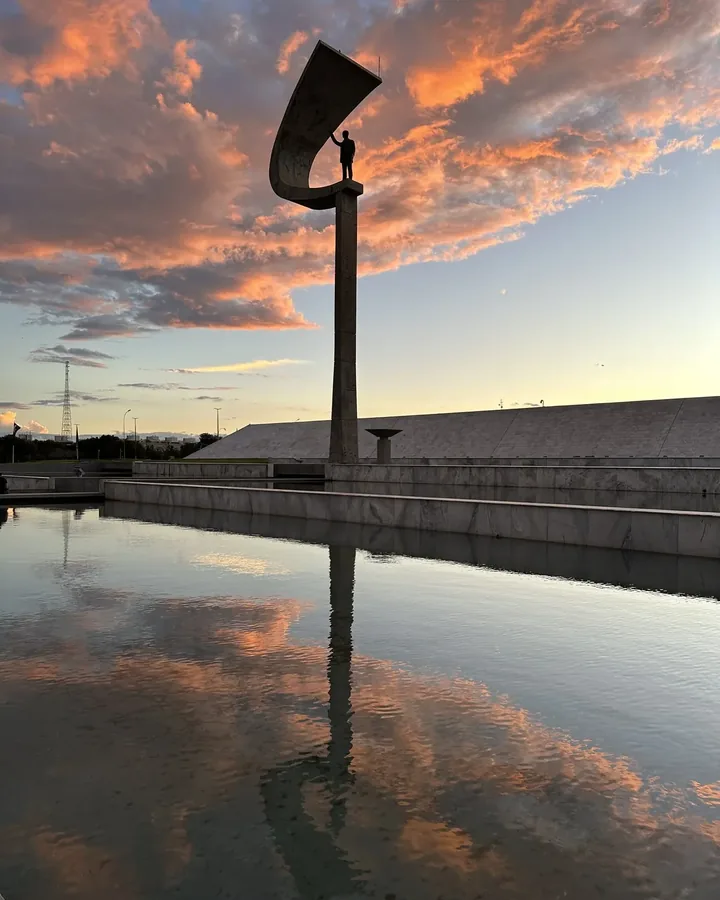 Memorial JK e bandeiras refletidos na água, sob céu azul e alaranjado.