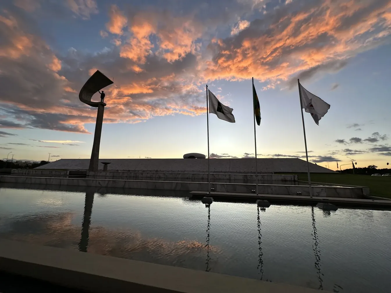 Memorial JK, bandeiras e céu azul e laranja refletidos no espelho d’água, fotografados com iPhone.