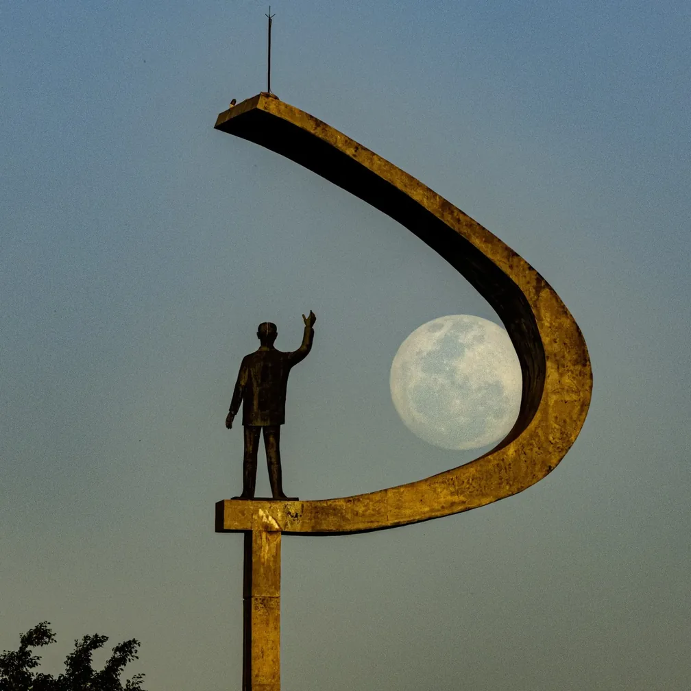 Lua grande e detalhada junto à mão da estátua de JK, cercada pelo arco do monumento.